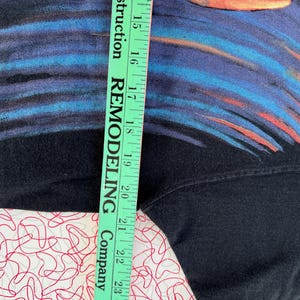 May include: Black t-shirt with a blue and orange abstract design. The shirt is being measured with a ruler that says "Remodeling Company" on it.