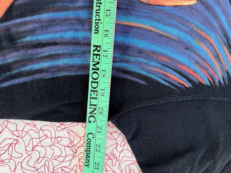 May include: Black t-shirt with a blue and orange abstract design. The shirt is being measured with a ruler that says "Remodeling Company" on it.