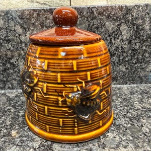 May include: A cylindrical ceramic honey pot with a glossy brown finish, featuring a textured basket-weave pattern and raised bee motifs on the exterior. The container includes a matching removable lid with a rounded knob handle on top.
