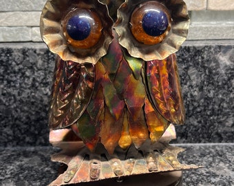 May include: A vintage metal owl music box with iridescent copper finish. The owl has large, round eyes with blue and orange glass accents. The music box has a wind-up mechanism and plays a tune.