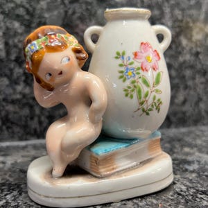 May include: A porcelain figurine of a girl with brown hair and a floral headband sitting on a book next to a white vase with floral designs.