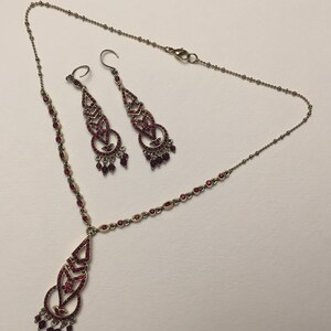 May include: A set of red crystal jewelry featuring a necklace and a pair of dangle earrings. The necklace and earrings have a delicate, intricate design with a Celtic knot motif.