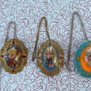 May include: Three vintage doll pendants with gold-tone chains. Each pendant features a small doll inside an oval frame. The frames are gold and turquoise, with decorative details. The dolls wear dresses.