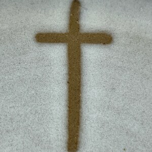 May include: A brown cross drawn in the sand on a white surface.