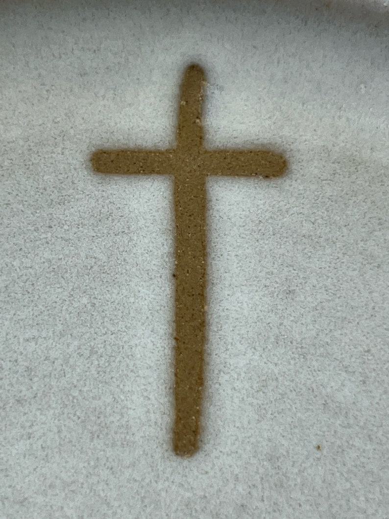 May include: A brown cross drawn in the sand on a white surface.
