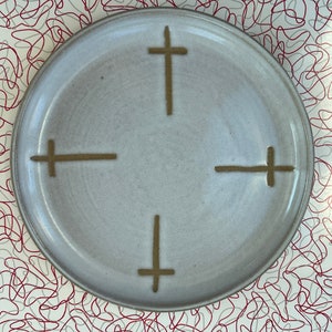 May include: A white ceramic plate with a brown cross design in the center. The plate has a slightly textured surface.