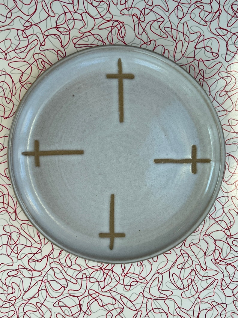 May include: A white ceramic plate with a brown cross design in the center. The plate has a slightly textured surface.