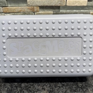 May include: A white plastic pencil box with a textured surface and the word "SpaceMaker" embossed on the lid. The box is rectangular with rounded corners and small, raised dots around the edges.