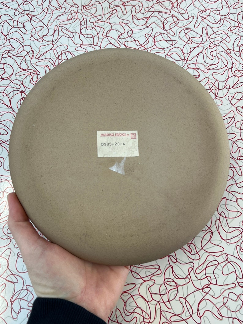 May include: A brown ceramic plate with a white label that reads "MARSHALL STUDIOS INC. DG85-28-4".