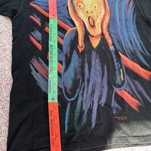 May include: Black t-shirt with a colorful graphic print of Edvard Munch's "The Scream". The print features a person with their hands over their ears, a bright yellow face, and a wide-open mouth. The background is a swirling mix of blue, green, and orange.