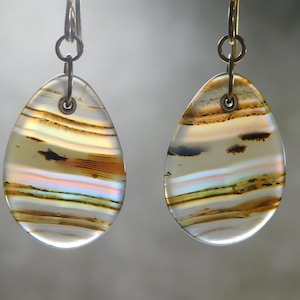 May include: A pair of teardrop-shaped earrings. Each earring features a translucent, light-colored stone with brown and gold striations. The earrings are suspended from gold-colored metal hooks, creating a natural and elegant accessory.