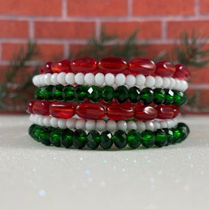 May include: A stack of six bracelets made with red, white, and green glass beads. The bracelets are stacked on a white surface.