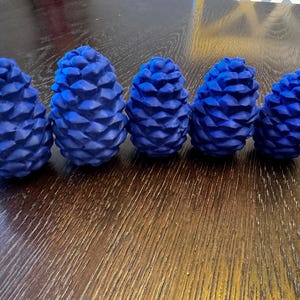 May include: Five blue pine cone decorations of varying sizes are arranged in a row on a wooden surface. The cones have a textured, geometric pattern and are a vibrant shade of blue. The wooden surface has a warm, brown tone.