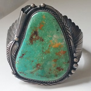 May include: A silver and turquoise Native American bracelet with a large, green turquoise stone set in a silver bezel. The bracelet is decorated with silver feathers and other designs.