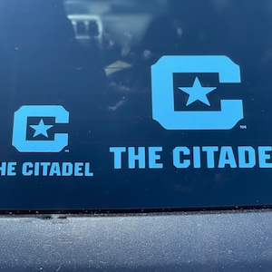 May include: Two blue and white decals on a black surface. The larger decal features a stylized "C" with a star inside and the text "THE CITADEL". The smaller decal is the same design but smaller.