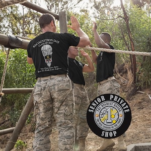 May include: Three people in black t-shirts and camouflage pants high-fiving outdoors. The shirts have text and a skull graphic. The text reads "RECOGNITION DAY CLASS OF 2027" and "The Pain Goes Away, But the Pride Lasts Forever."