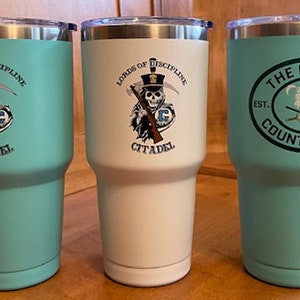 May include: Three stainless steel tumblers with different designs. The first tumbler is teal and features a skull and crossbones with the text "Lords of Discipline The Citadel". The second tumbler is white and features the same skull and crossbones design. The third tumbler is teal and features a green circle with the text "The Citadel Country Club Est. 1946".