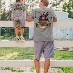 May include: Two people wearing grey t-shirts with a graphic print of a shield with the text "Our Citadel" and a depiction of a military unit. The shield also includes the text "History Binds Us, Tradition Keeps Us Strong".