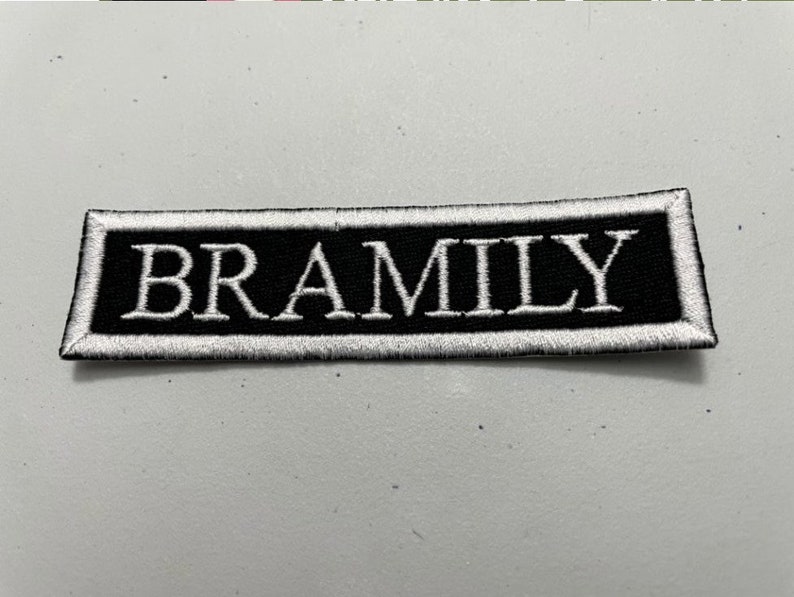 以下が含まれることがあります： 黒い背景に白い大文字でBRAMILYという単語が刺繍された長方形のワッペン。ワッペンには太い白い縁取りがあり、端はきれいに縫製されており、コントラストの強いデザインに仕上がっています。