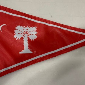 May include: A red triangular flag with a white border and a white image of a palmetto tree and crescent moon. The flag is made of fabric and has a white backing.