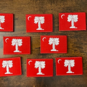May include: Rectangular red patches featuring the South Carolina state flag design. Each patch displays a white palmetto tree and a crescent moon. The patches are embroidered with white thread and have a black border.