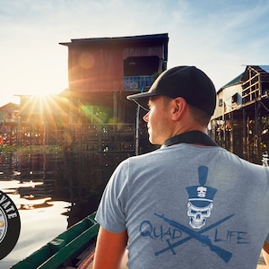 May include: A light gray t-shirt with a skull and crossed rifles design, and the words "QUAD LIFE". The image also includes a black baseball cap and a shop logo. The background shows a river and buildings.