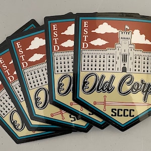 May include: Five black and teal stickers with a beige background. The stickers feature a gray castle with a red roof and white clouds. The text "Old Corps" is in black cursive lettering. The text "ESTD 1842" is in black lettering. The text "SCCC" is in black lettering.