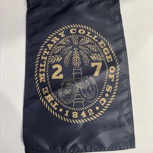 May include: A rectangular navy blue flag featuring a gold circular emblem. The text reads THE MILITARY COLLEGE OF S.C. 1842, with a large palm tree, the number 27, and the South Carolina state seal in the center. The design is printed in gold ink.