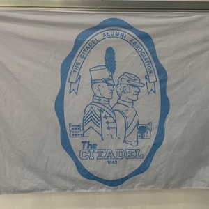 May include: A white flag with a blue oval containing the text "The Citadel Alumni Association 1842" and a graphic of two men in uniform.
