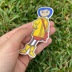 May include: A sticker featuring a cartoon character with blue hair, wearing a yellow raincoat, red dress, and yellow boots. The character is carrying a red bag. The sticker is held in a hand against a background of green grass.