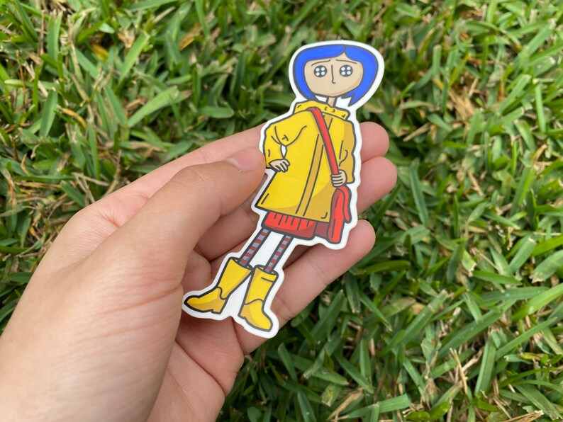 May include: A sticker featuring a cartoon character with blue hair, wearing a yellow raincoat, red dress, and yellow boots. The character is carrying a red bag. The sticker is held in a hand against a background of green grass.