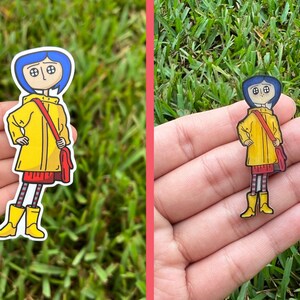 May include: A sticker featuring a cartoon character with blue hair, wearing a yellow raincoat, red skirt, and boots. The character has a red bag slung over their shoulder. The sticker is set against a background of green grass.