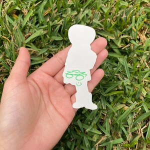 May include: A white die-cut sticker in the shape of a person, held in a hand against a backdrop of green grass. The sticker features green scribbled details and a smiling face.