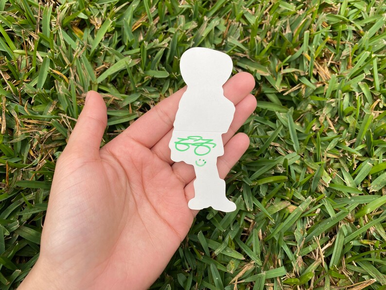May include: A white die-cut sticker in the shape of a person, held in a hand against a backdrop of green grass. The sticker features green scribbled details and a smiling face.