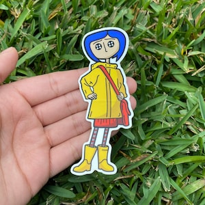 May include: A sticker featuring a cartoon character with blue hair, button eyes, and a yellow raincoat. The character is holding a red bag and wearing a red skirt, striped socks, and yellow boots. The sticker is on a white background.