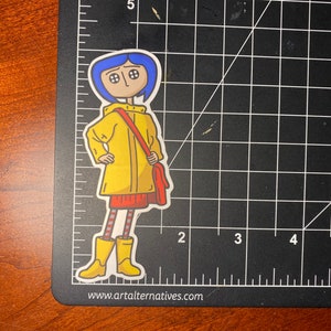 May include: A die-cut sticker featuring a cartoon character with blue hair, button eyes, and a yellow raincoat. The character is wearing a red skirt, striped leggings, and yellow boots, carrying a red bag. The sticker is on a black cutting mat.