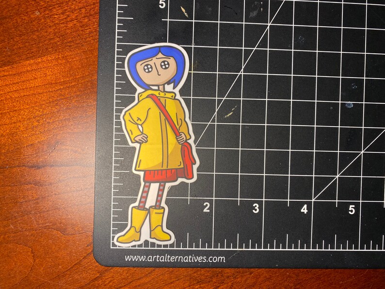 May include: A die-cut sticker featuring a cartoon character with blue hair, button eyes, and a yellow raincoat. The character is wearing a red skirt, striped leggings, and yellow boots, carrying a red bag. The sticker is on a black cutting mat.