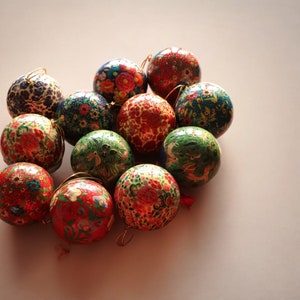 Può includere: Una collezione di 12 addobbi natalizi dipinti a mano in vari colori e motivi, tra cui rosso, verde, blu e oro. Gli addobbi sono realizzati in cartapesta e presentano intricati disegni floreali.