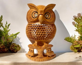 May include: A wooden owl sculpture with intricate details. The owl is carved with a detailed pattern and has large, round eyes. The owl is perched on a tree stump with two smaller owls on either side.
