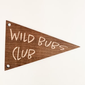 May include: A wooden pennant flag with the words "WILD BUBS CLUB" in a handwritten style. The pennant is a dark brown color and has two silver rivets in the corners. The background is white.
