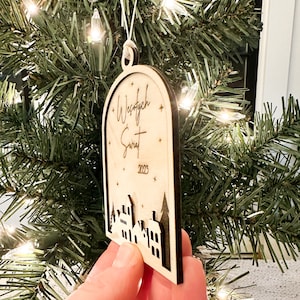 May include: A wooden Christmas ornament with a white background and black lettering. The ornament features the text "Wesołych Świąt" and the year "2023". The ornament also features a silhouette of a small town with houses and trees.