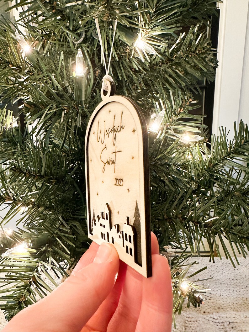 May include: A wooden Christmas ornament with a white background and black lettering. The ornament features the text "Wesołych Świąt" and the year "2023". The ornament also features a silhouette of a small town with houses and trees.