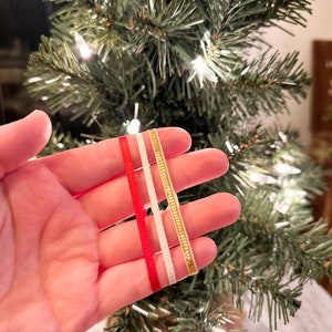 May include: Three different ribbons in red, white, and gold. The ribbons are held in a hand in front of a small Christmas tree.