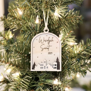May include: A wooden Christmas ornament with a white background and brown text. The ornament features a small town silhouette and the text "Wesołych Świąt 2023".