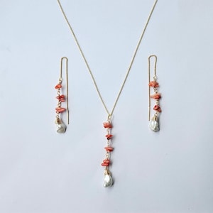 Op de afbeelding: Een goudkleurige ketting en een paar bijpassende doorrijkoorbellen. De ketting heeft een centraal hangertje met kleine, onregelmatige koraalkleurige steensplinters en een witte barokparel aan het uiteinde. Elke oorbel is identiek.