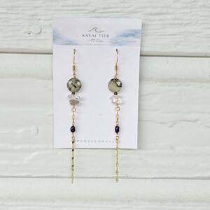 May include: Dangle earrings on a white display card labeled Kauai Tide Atelier. Each earring features a faceted sage green bead, a clear crystal, a small black bead, and a long gold-tone chain. The design is symmetrical with gold-tone hooks.