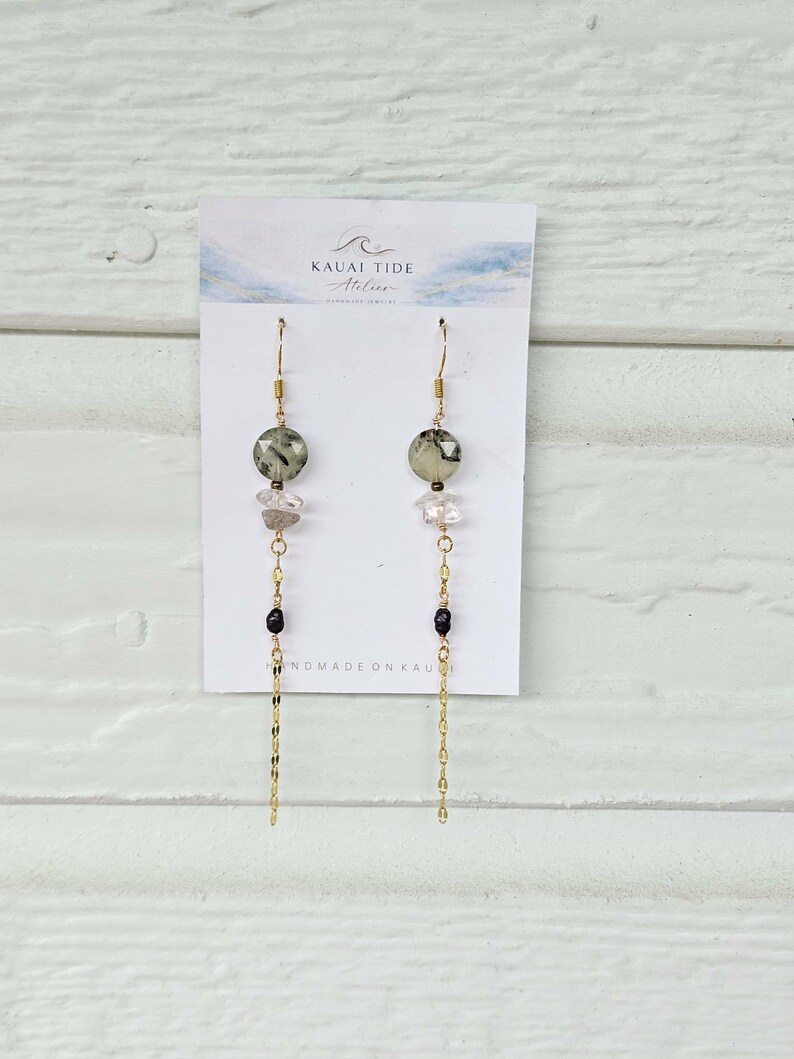 May include: Dangle earrings on a white display card labeled Kauai Tide Atelier. Each earring features a faceted sage green bead, a clear crystal, a small black bead, and a long gold-tone chain. The design is symmetrical with gold-tone hooks.