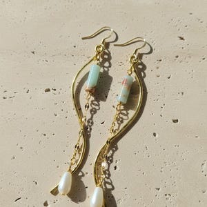 May include: A pair of gold-toned dangle earrings featuring a curved metal wire, a delicate chain, a cylindrical light green stone bead with brown accents, and a single white teardrop-shaped pearl at the base, suspended from gold-toned ear hooks.