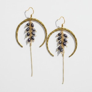 May include: A pair of gold-toned earrings featuring a large, hammered crescent-shaped metal frame. Suspended from the center of each crescent is a cluster of small, dark grey oval beads attached to delicate gold chains that hang downward.