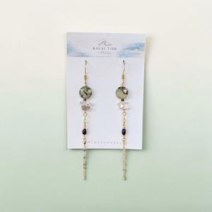 May include: A pair of gold-tone dangle earrings on a white display card labeled Kauai Tide Atelier. Each earring features a faceted sage green bead, a clear crystal chip, a small dark bead, and a delicate gold-tone chain hanging from a hook.